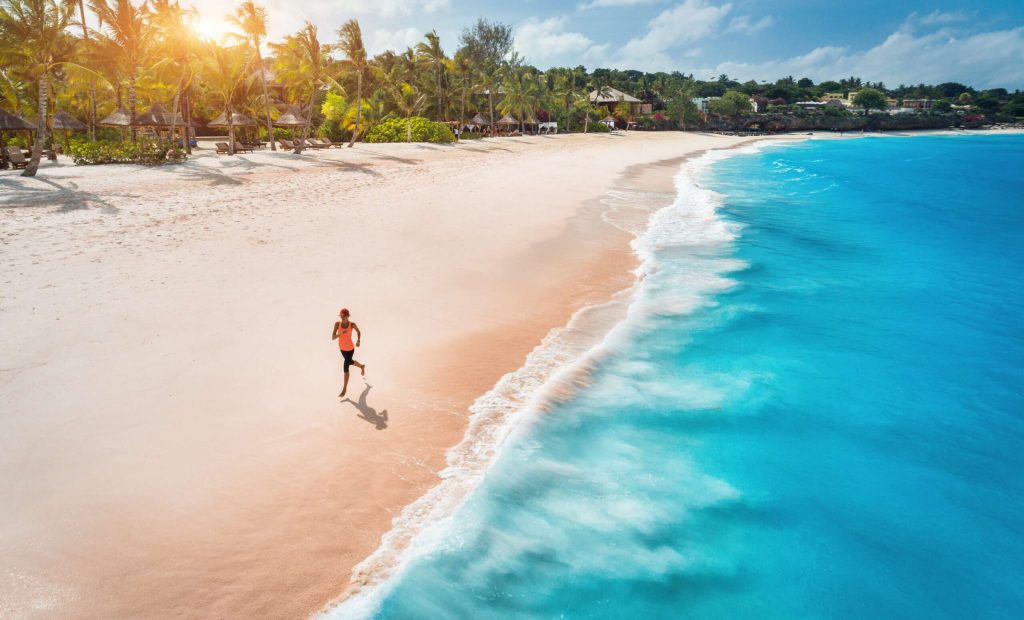 Aerial view of the running young woman on the white sandy beach near sea with waves at sunset. Summer holiday. Top view of sporty slim girl, clear azure water. Indian Ocean. Lifestyle and sport