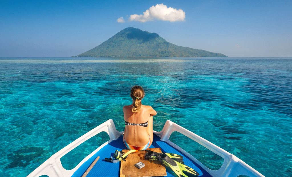Young woman in swimsuit sit on boat with a mask and flippers looking to a clean sea and volcano Manado Tua. North Sulawesi, Indonesia.